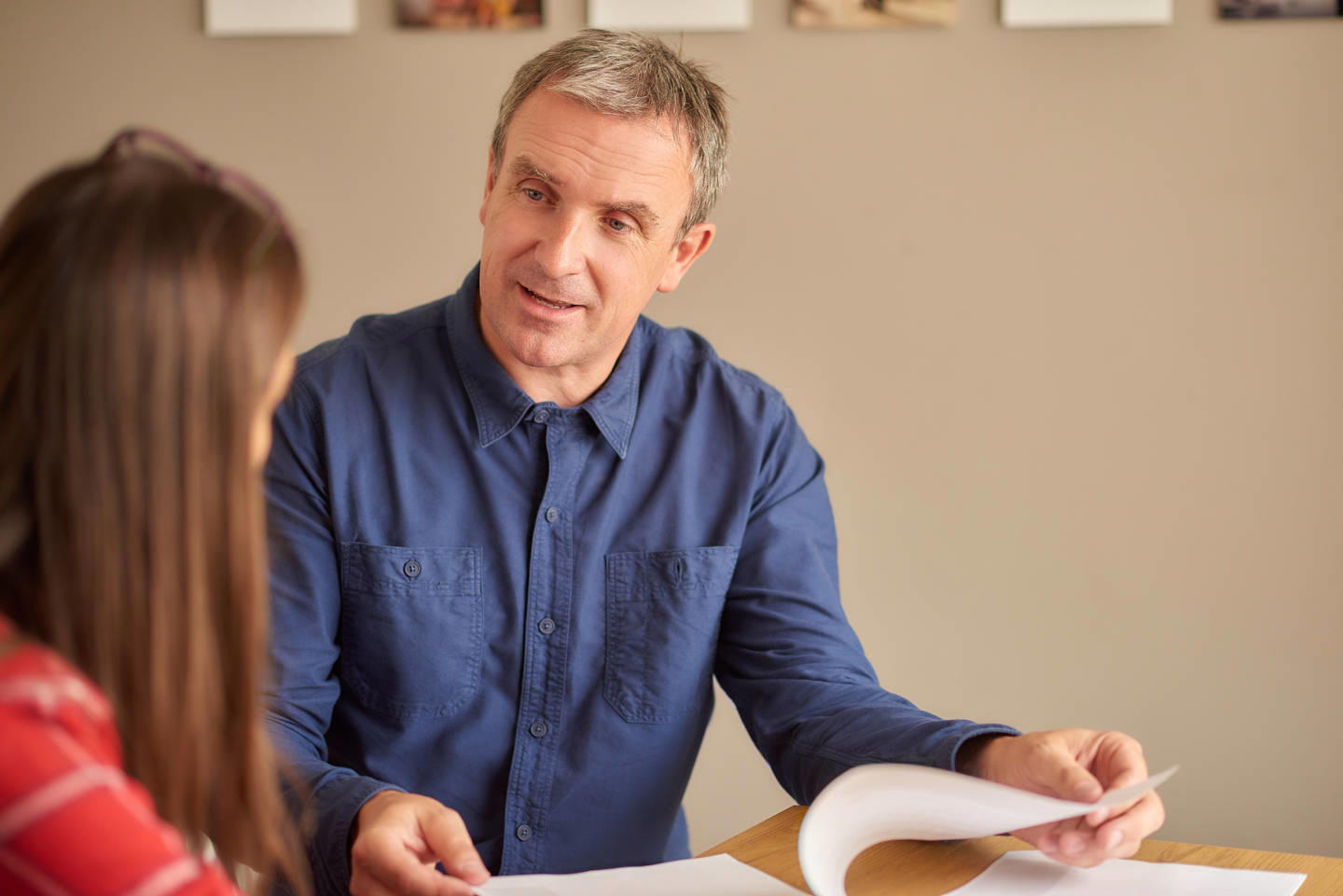 A man sits speaking to a woman with her back to the camera. He is holding open a stapled document.
