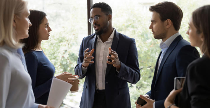 People in business attire stood in a tight circle in front of a window. A man is explaining something and gesticulating with his hands