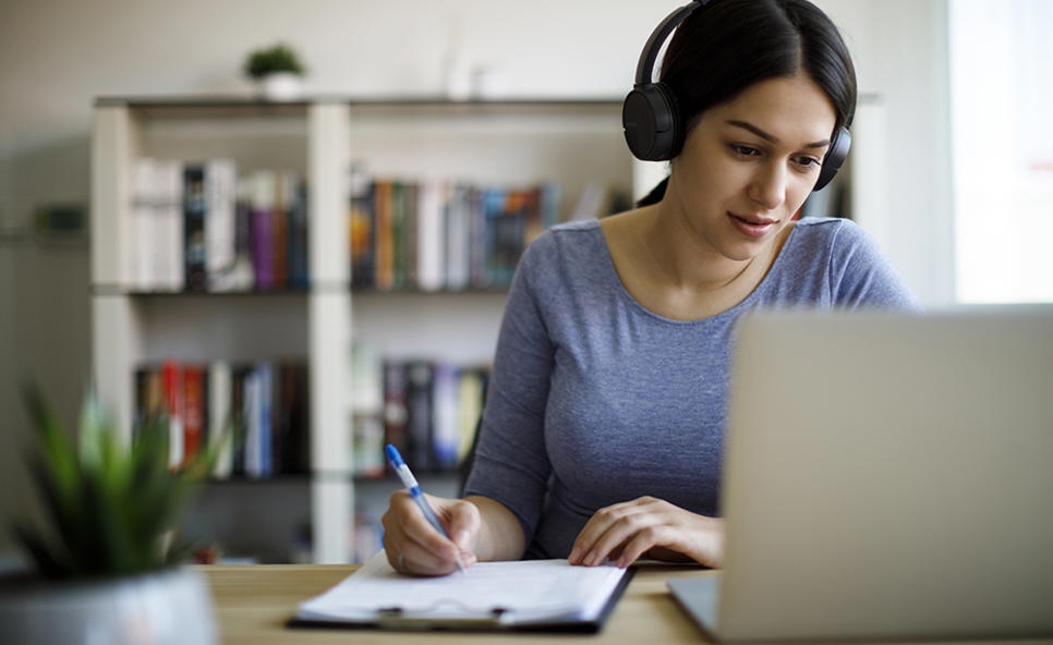 A woman in headphones sits at a desk in the library. She is writing in her notebook and paying attention to something on her laptop screen.