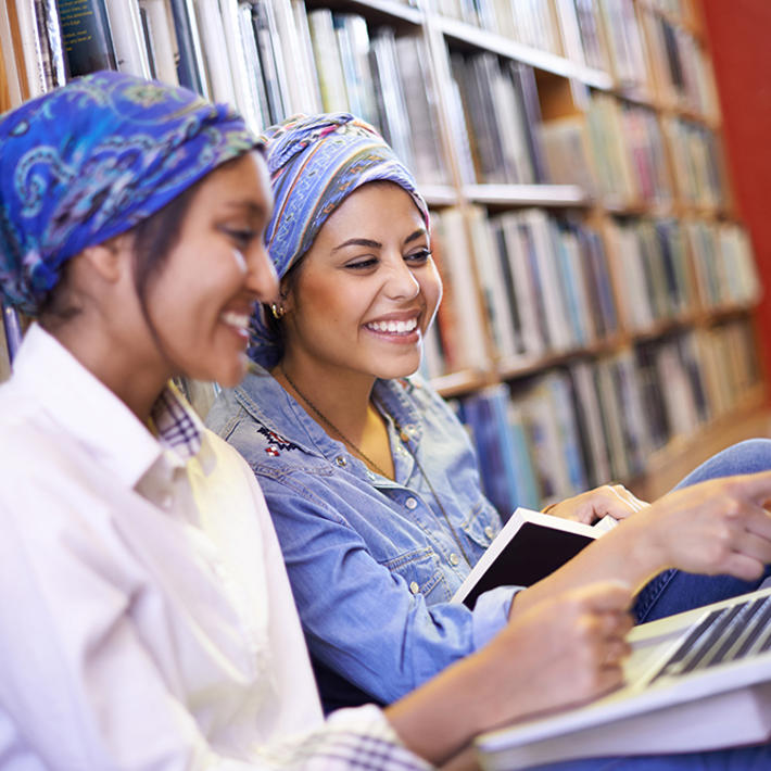 Two women smiling and sat on the floor leaning against a bookshelf with laptops open