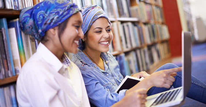 Two women smiling and sat on the floor leaning against a bookshelf with laptops open
