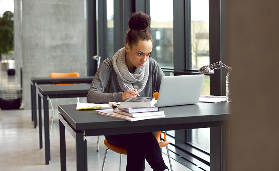 A woman sat alone at a library table near a window in front of books and laptop. She is concentrating on her screen with a pen in hand.