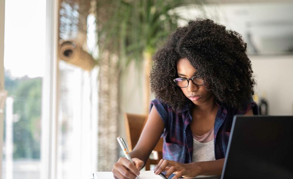 A woman sat at a desk by a window, writing in her notebook. Her laptop is open in the foreground.