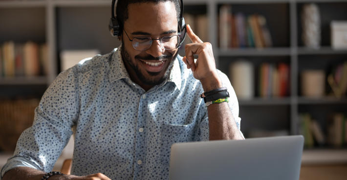 A smiling man wearing a headset in conversation at his laptop. A metal framed bookshelf is in the background and out of focus.